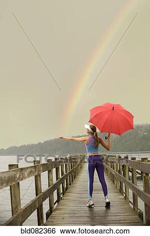 Stock Photograph - Caucasian woman in sportswear with red umbrella on pier catching rainbow. Fotosearch