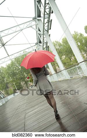 Stock Image - Caucasian woman walking in rain with red umbrella. Fotosearch