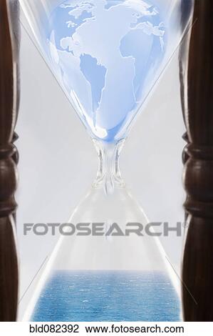 Stock Image - Globe squeezing through hour glass. Fotosearch
