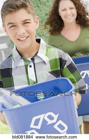 Picture - Hispanic boy carrying recycling bin with mother in background. Fotosearch