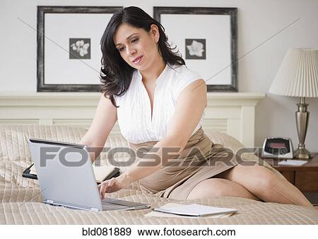 Stock Photo - Hispanic businesswoman using laptop on bed in bedroom. Fotosearch