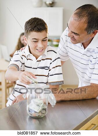 Stock Photo - Hispanic father watching son put money into jar. Fotosearch