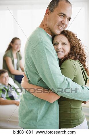 Stock Image - Hispanic husband and wife hugging with family in background. Fotosearch
