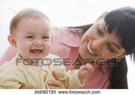 Stock Image - Hispanic mother watching laughing baby girl. Fotosearch