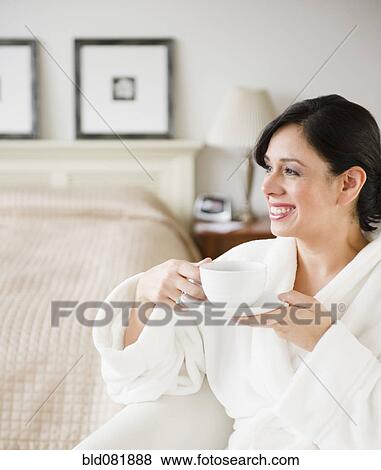 Stock Photo - Hispanic woman in bathrobe drinking coffee in bedroom. Fotosearch