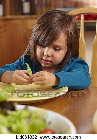 Stock Image - Mixed race girl shelling pea pod at dining table. Fotosearch