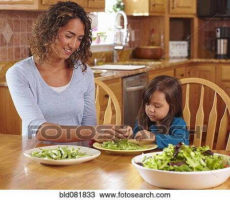 Stock Image - Mother and daughter shelling peas at dining table. Fotosearch