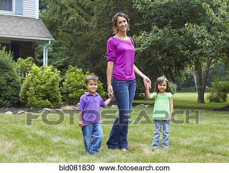 Stock Image - Mother holding hands with son and daughter in front yard. Fotosearch