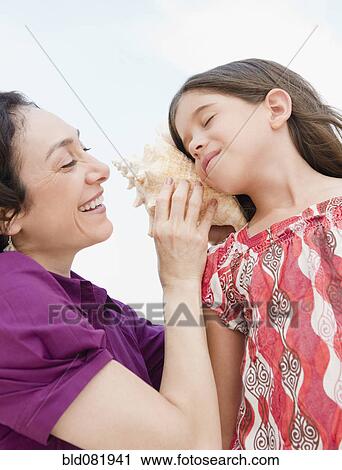 Stock Image - Mother holding seashell to daughter's ear. Fotosearch