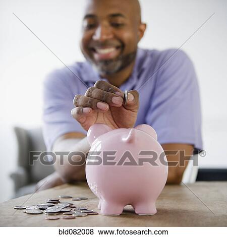 Stock Photo - Smiling African American man putting coins in piggy bank. Fotosearch