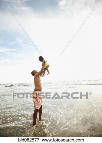 Stock Photo - Black father lifting daughter while standing in ocean. Fotosearch