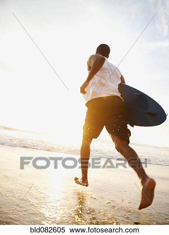 Stock Photography - Black man running with surfboard on beach. Fotosearch