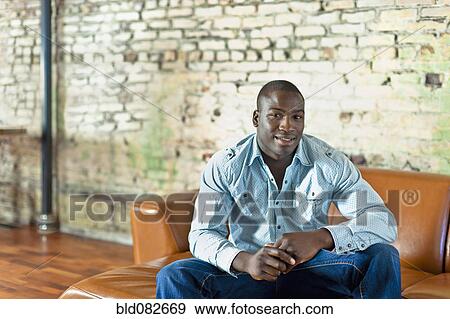 Black man sitting on sofa in living room View Large Photo Image Stock Photo - Black man sitting on sofa in living room. Fotosearch