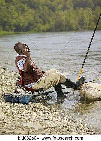 Picture - Black man with cell phone fishing in stream. Fotosearch