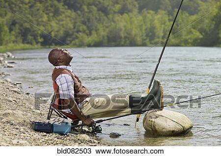 Stock Image - Black man with feet up fishing in stream. Fotosearch