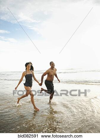 Stock Image - Couple holding hands and running on beach. Fotosearch