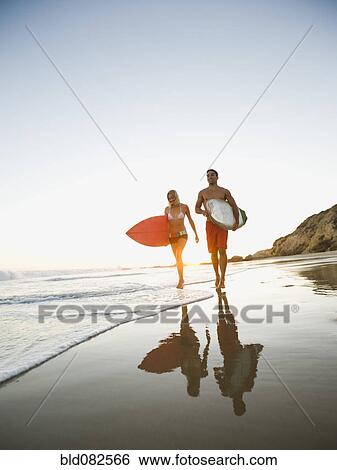 Stock Photograph - Couple walking on beach carrying surfboards. Fotosearch