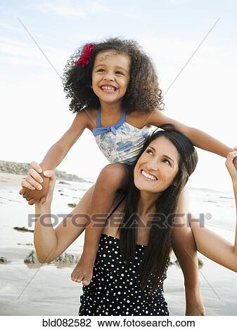 Stock Image - Hispanic mother carrying daughter on shoulders at beach. Fotosearch