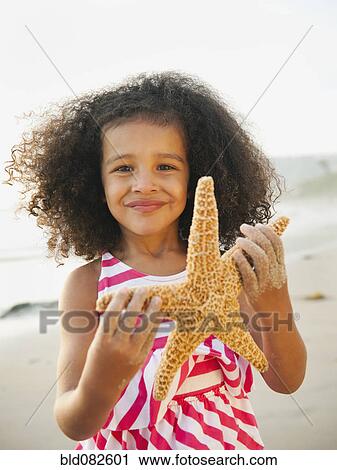 Stock Image - Mixed race girl holding starfish on beach. Fotosearch