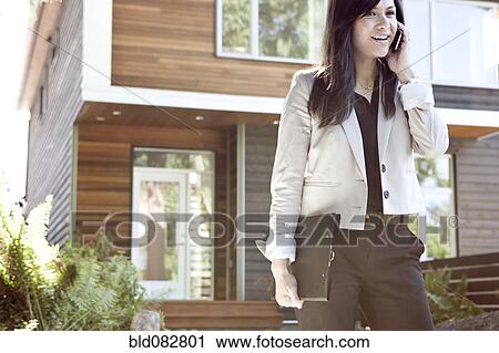 Stock Image - Mixed race real estate agent standing near house talking on cell phone. Fotosearch