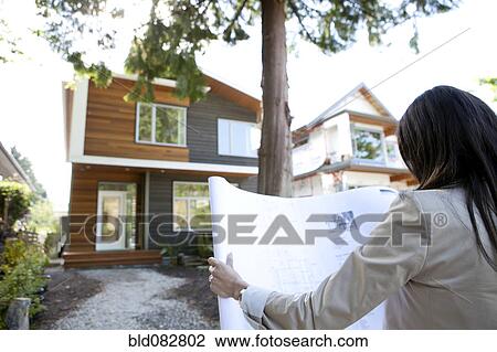 Stock Image - Mixed race real estate agent standing near house reviewing blueprints. Fotosearch