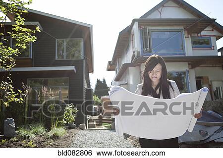 Stock Photograph - Mixed race real estate agent standing near house reviewing blueprints. Fotosearch
