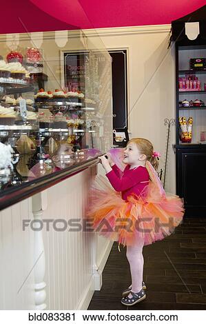 Stock Image - Caucasian girl in tutu looking at cupcakes in bakery. Fotosearch