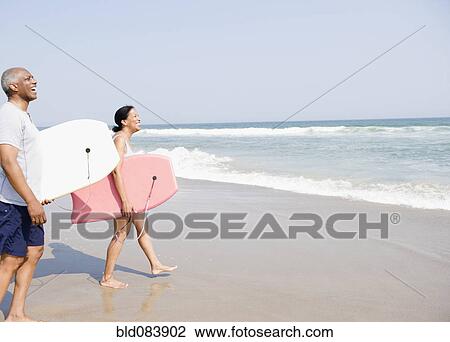 Stock Image - Black couple holding body boards on beach. Fotosearch