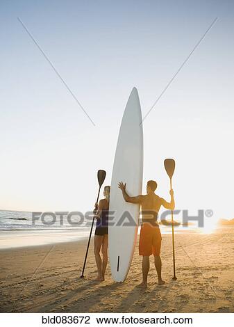 Stock Image - Couple standing on beach with surfboard and paddles. Fotosearch