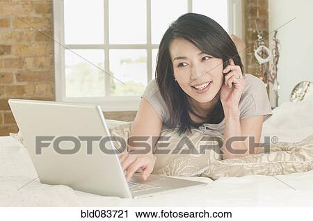 Stock Image - Korean woman laying on bed using laptop and talking on phone. Fotosearch