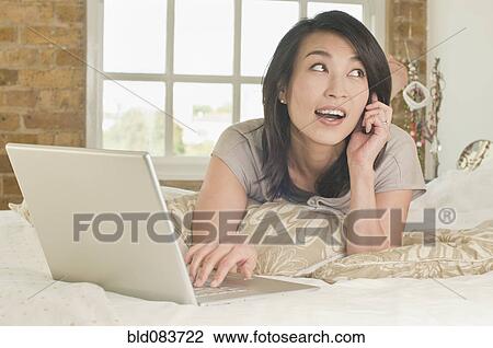 Stock Image - Korean woman laying on bed using laptop and talking on phone. Fotosearch