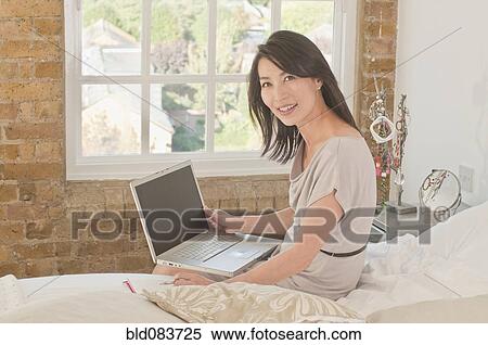 Stock Photography - Korean woman sitting on bed using laptop. Fotosearch