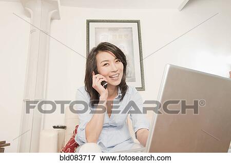 Stock Photo - Korean woman using laptop and talking on phone in living room. Fotosearch