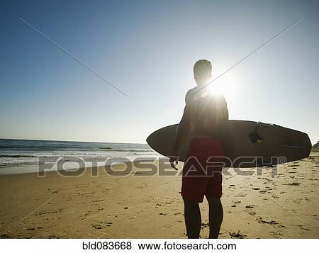 Stock Photo - Mixed race man carrying surfboard on beach. Fotosearch