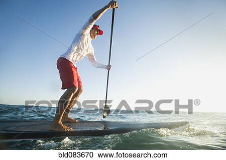 Stock Image - Mixed race man paddling on surfboard. Fotosearch