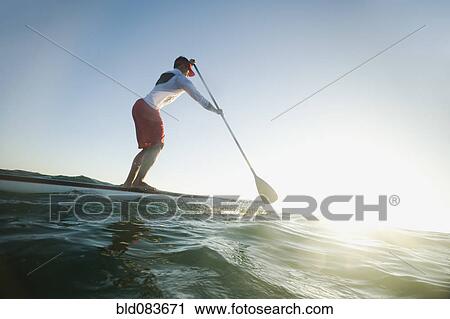 Stock Image - Mixed race man paddling on surfboard. Fotosearch