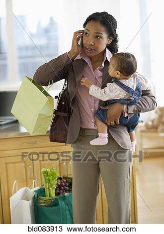 Stock Image - Mixed race mother holding baby and talking on cell phone. Fotosearch
