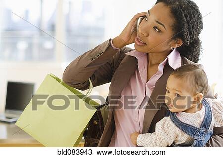 Stock Image - Mixed race woman holding baby and talking on cell phone. Fotosearch