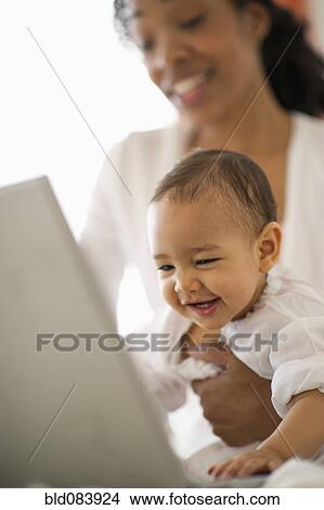Picture - Mixed race woman holding baby and using laptop. Fotosearch