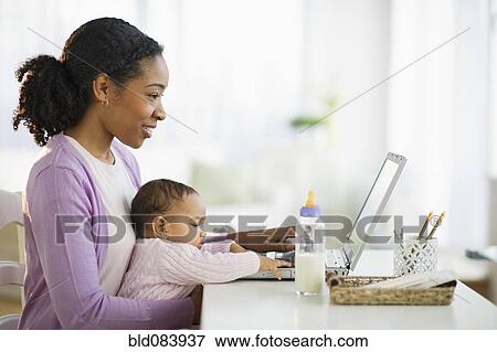 Stock Photo - Mixed race woman holding baby and using laptop. Fotosearch