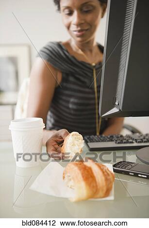 Stock Image - Black businesswoman eating croissant at desk. Fotosearch