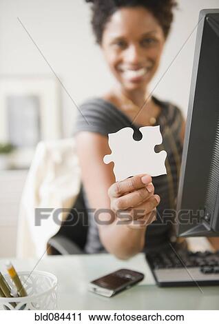 Stock Image - Black businesswoman holding puzzle piece at desk. Fotosearch