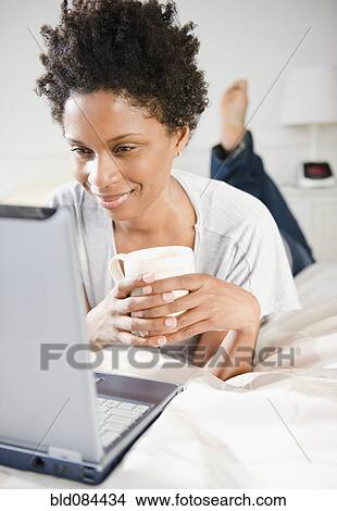 Picture - Black woman laying on bed using computer. Fotosearch