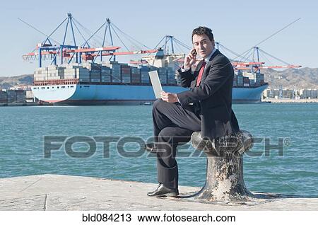 Stock Image - Hispanic businessman holding laptop with container ship in background. Fotosearch