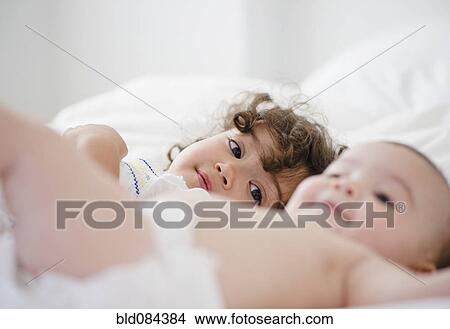 Picture - Mixed race girl watching baby sister on bed. Fotosearch