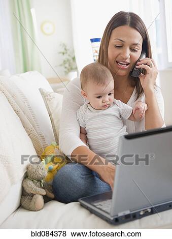 Stock Photo - Mother holding baby and using telephone and laptop. Fotosearch