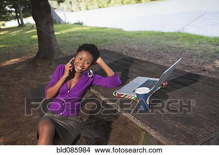 Picture - African American businesswoman sitting at picnic bench talking on cell phone. Fotosearch
