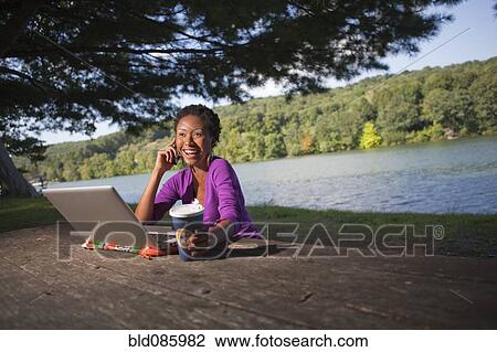 Stock Image - African American businesswoman sitting at picnic bench talking on cell phone. Fotosearch