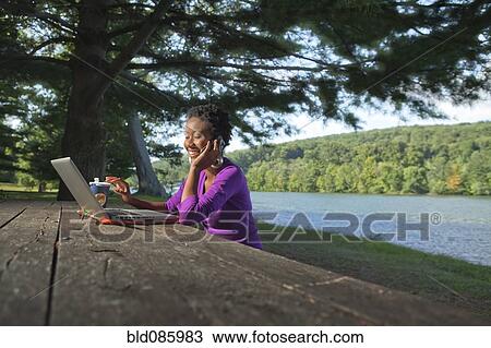 Stock Image - African American businesswoman sitting at picnic bench talking on cell phone. Fotosearch