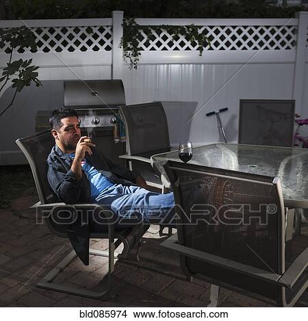 Picture - Caucasian man drinking wine and smoking on patio. Fotosearch